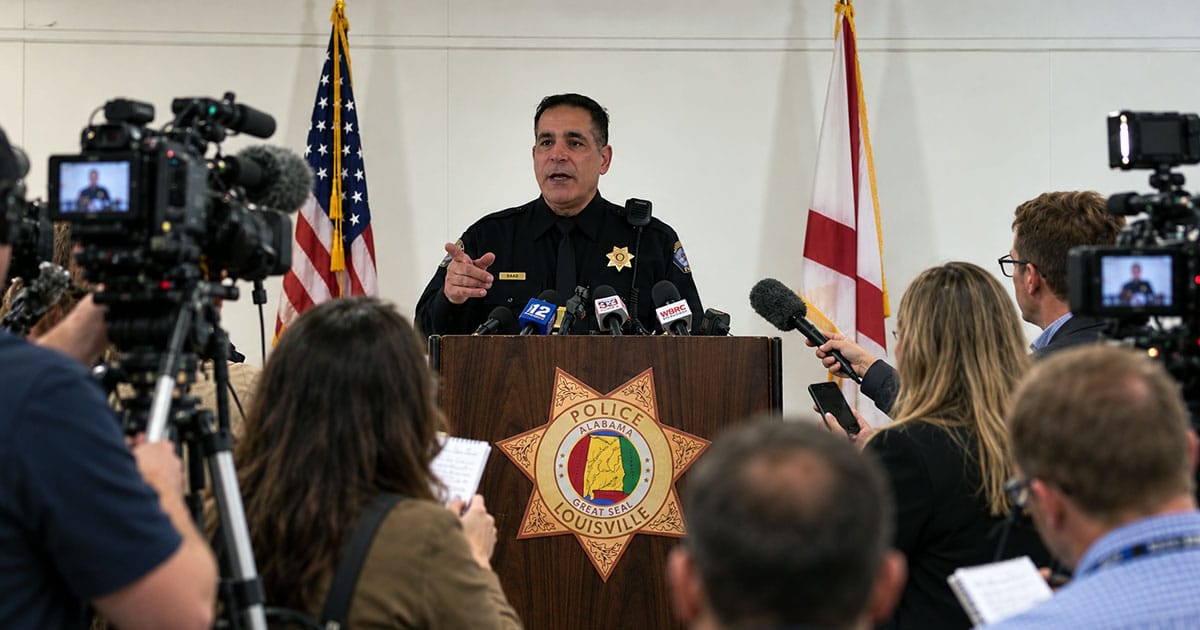 Police chief speaking at a podium while reporters, cameras, and microphones surround him during a press conference.