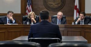Police chief candidate in a business suit facing a hiring board during an interview in city council chambers.