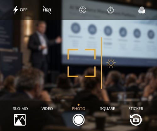 Phone-camera-style view of a corporate leadership summit, showing a speaker presenting an AI workforce management rollout to an audience.